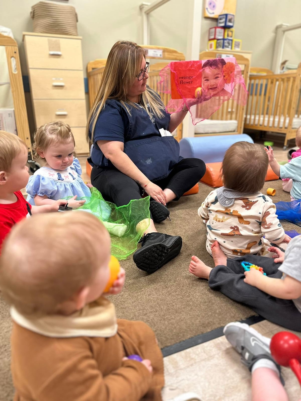 teacher sitting on a carpet reading a book to five babies