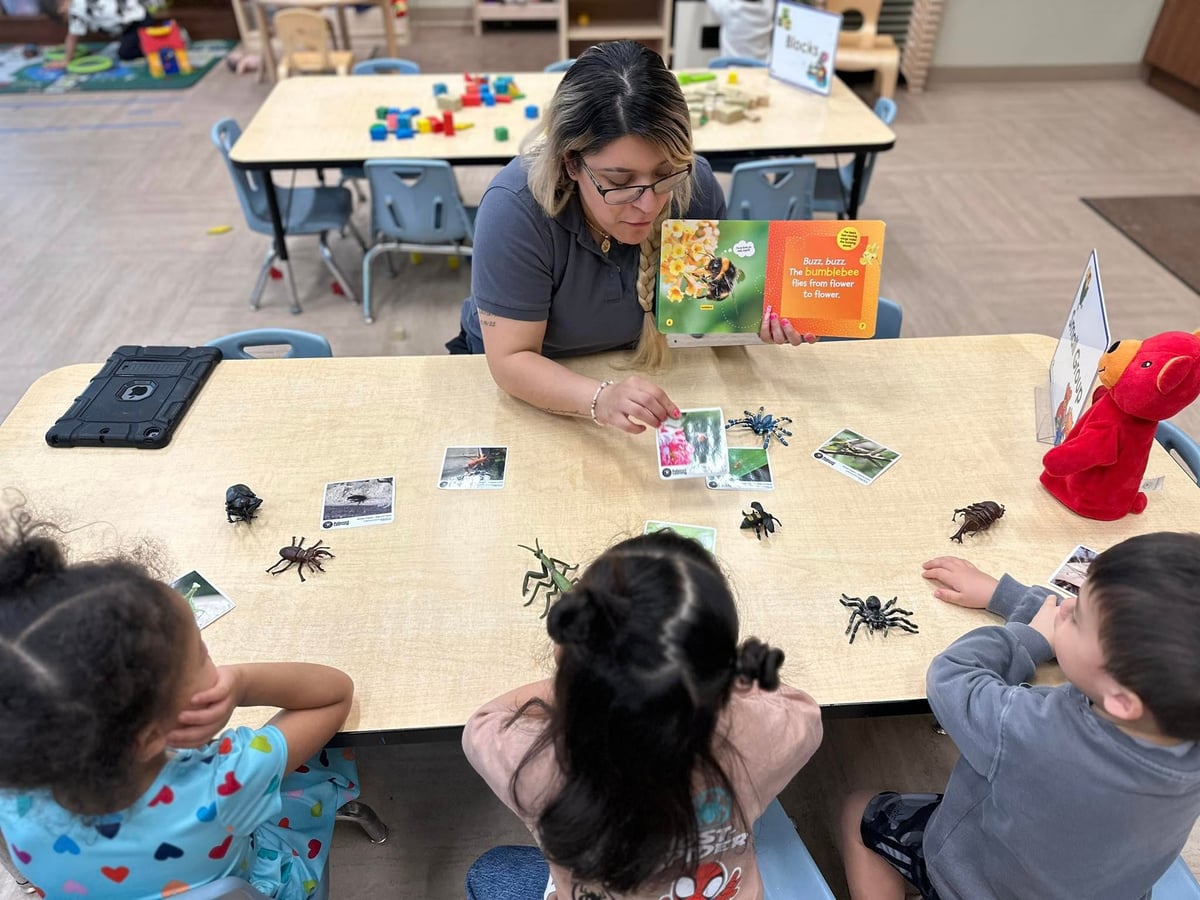 a teacher reading a book about insects to three children who are also observing toy insects on a table