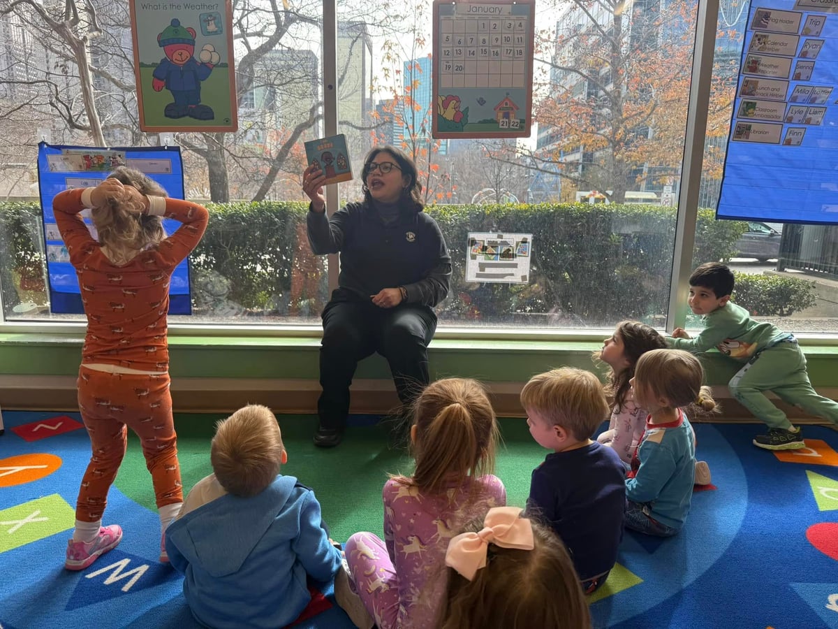 a teacher sitting in front of large windows, reading a book to eight children sitting on a colorful carpet