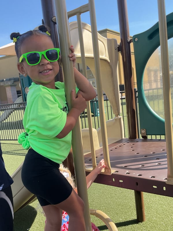 Preschool student wearing neon green clothing and sunglasses while climbing playground equipment during outdoor play time.