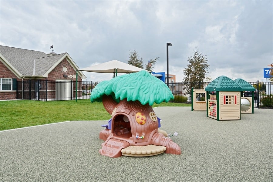 Toddler playground at Primrose School of Eagan featuring a whimsical tree house play structure, activity panels, artificial turf, and rubber safety surfacing with the school building in the background