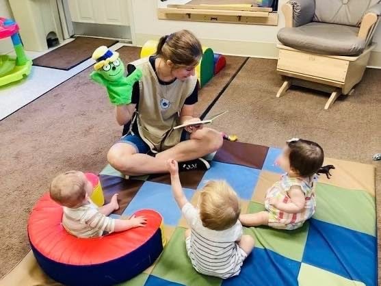 Infant teacher sitting on the floor with three children sitting around her while reading a book with an Og puppet on her hand.
