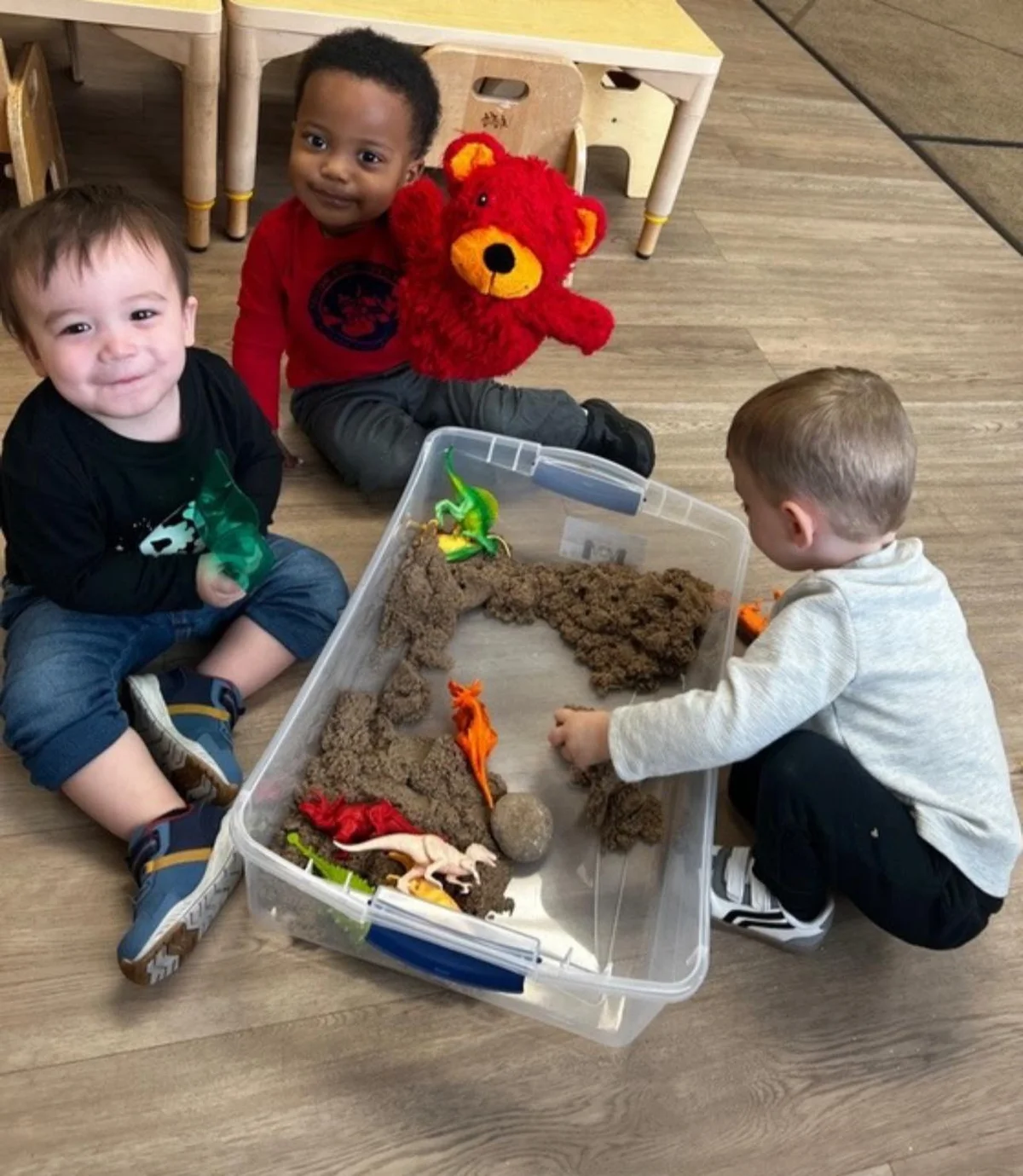 three toddlers smiling and playing with toy dinosaurs in a sensory bin