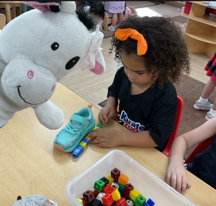 Preschool student using connecting cubes to measure the length of a shoe during a math and problem-solving activity with a classroom puppet nearby.