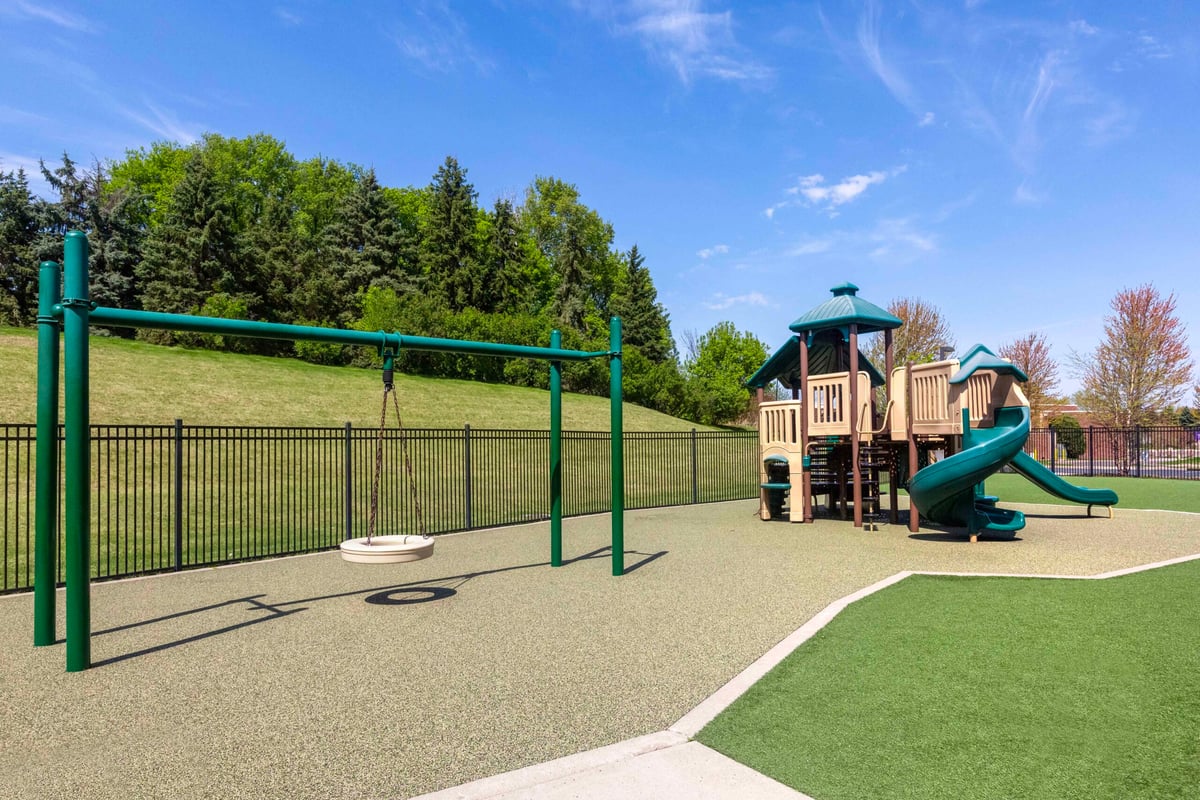 Outdoor playground at Primrose School of Eagan featuring a climbing structure, spiral slide, tire swing, and rubber safety surfacing, with a fenced perimeter and tree-lined hillside in the background.