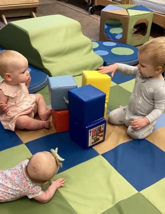 three babies on a colorful playmat, playing with large blocks