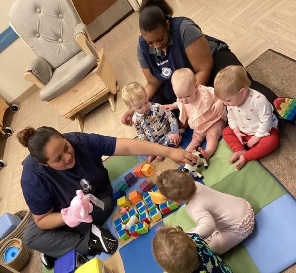 five babies engaging with two teachers, building blocks, and puppets