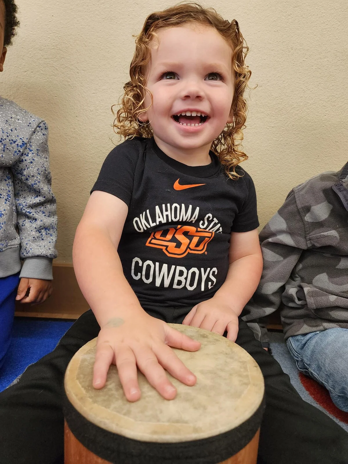 Toddler boy smiling while sitting on the carpet, beating the drum to the beat in a song during music time