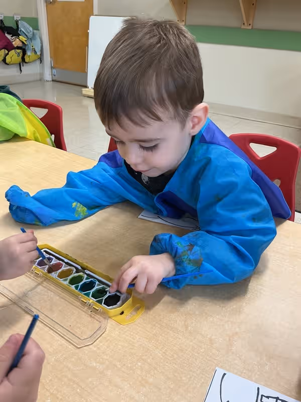 A boy in a smock painting with watercolors at a table.