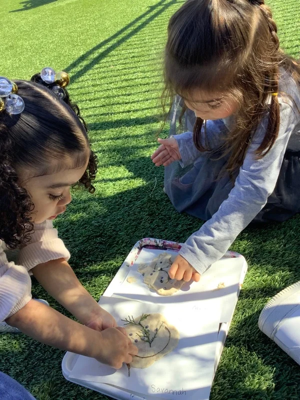 Two preschool girls engaging in an outdoor sensory activity, using natural materials to create faces on playdough, promoting creativity and fine motor development.