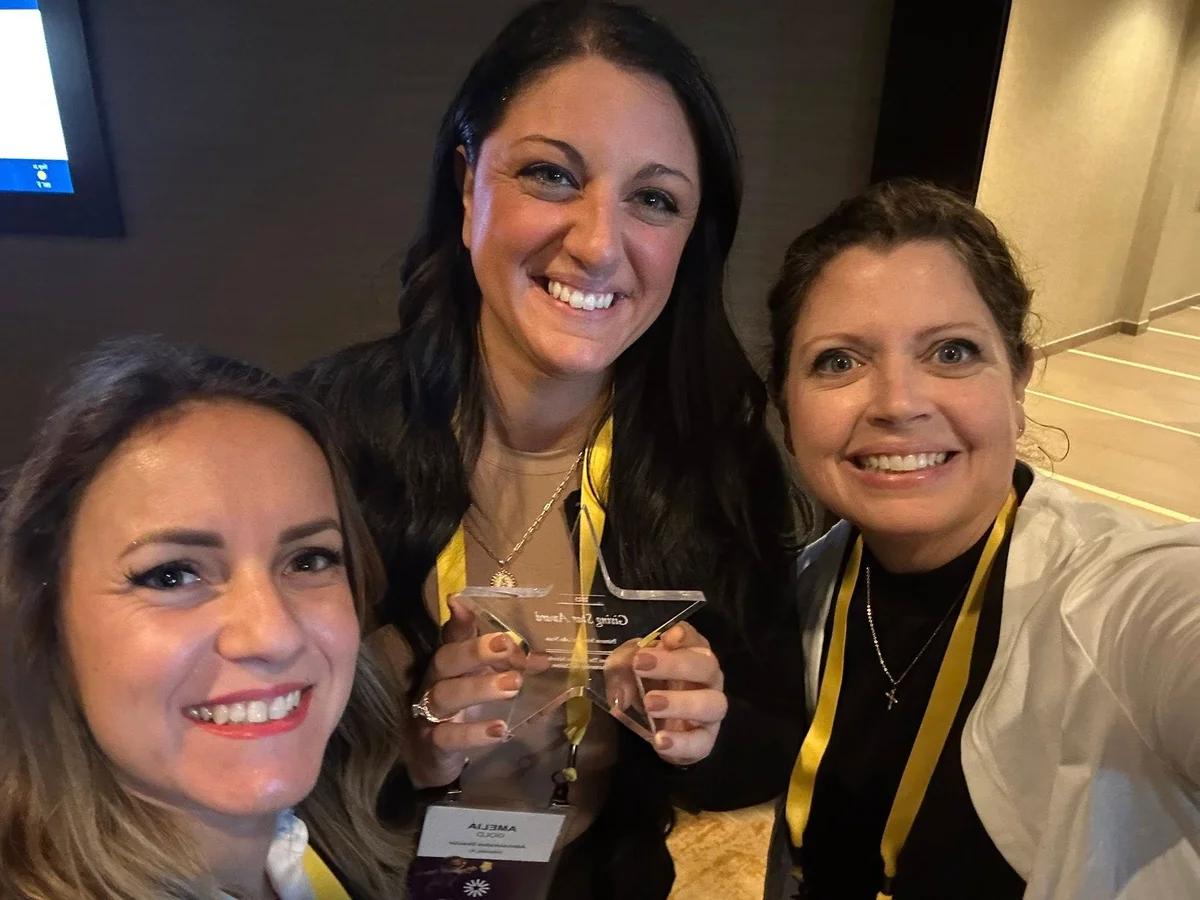 Three women smiling one with blonde hair two with brown hair wearing yellow lanyards holding a clear star award