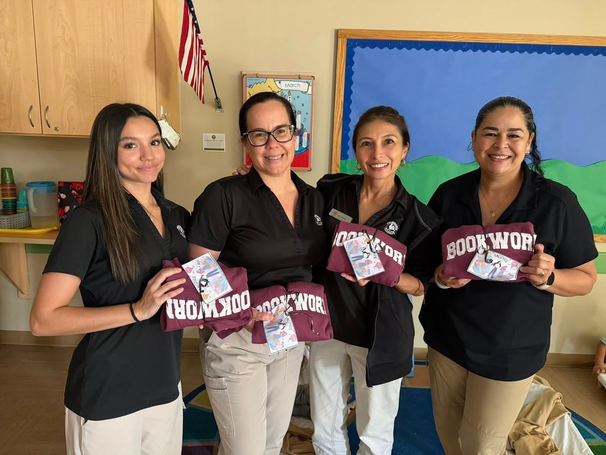four women in black polos and khaki pants holding a maroon shirt that says book worm in a classroom
