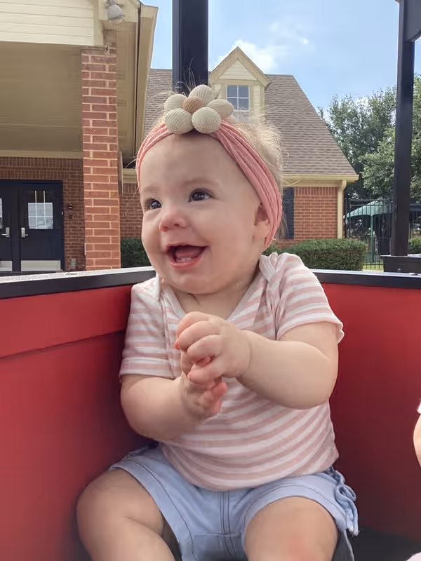 A baby girl with a pink flower bow riding in a red train car, with a happy smile on her face.