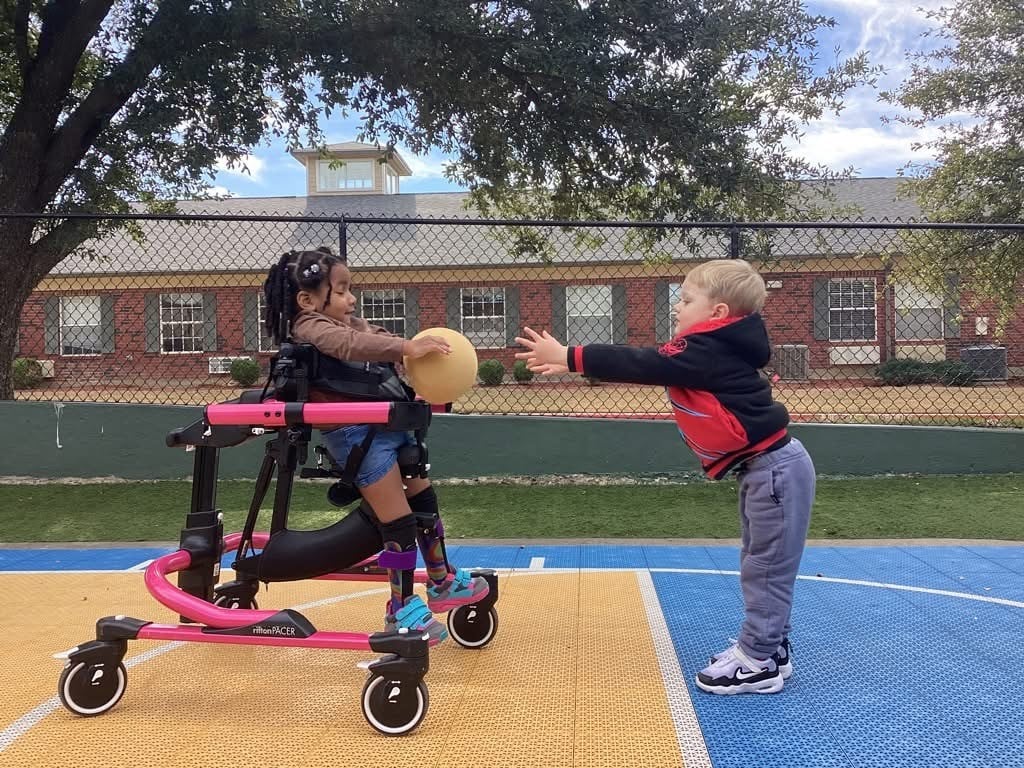 little girl in a wheeled walker tossing a ball with a friend on the playground