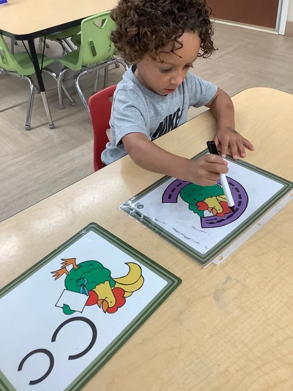 Preschool boy tracing the letter C on a laminated activity sheet with a dry erase marker, practicing early literacy and fine motor skills at a classroom table at the best childcare center in Fort Worth, TX.