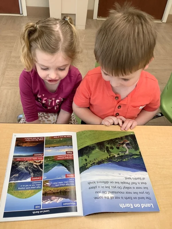 Two preschool children sitting side by side and reading a nonfiction book about landforms, building early literacy skills and exploring earth science concepts.