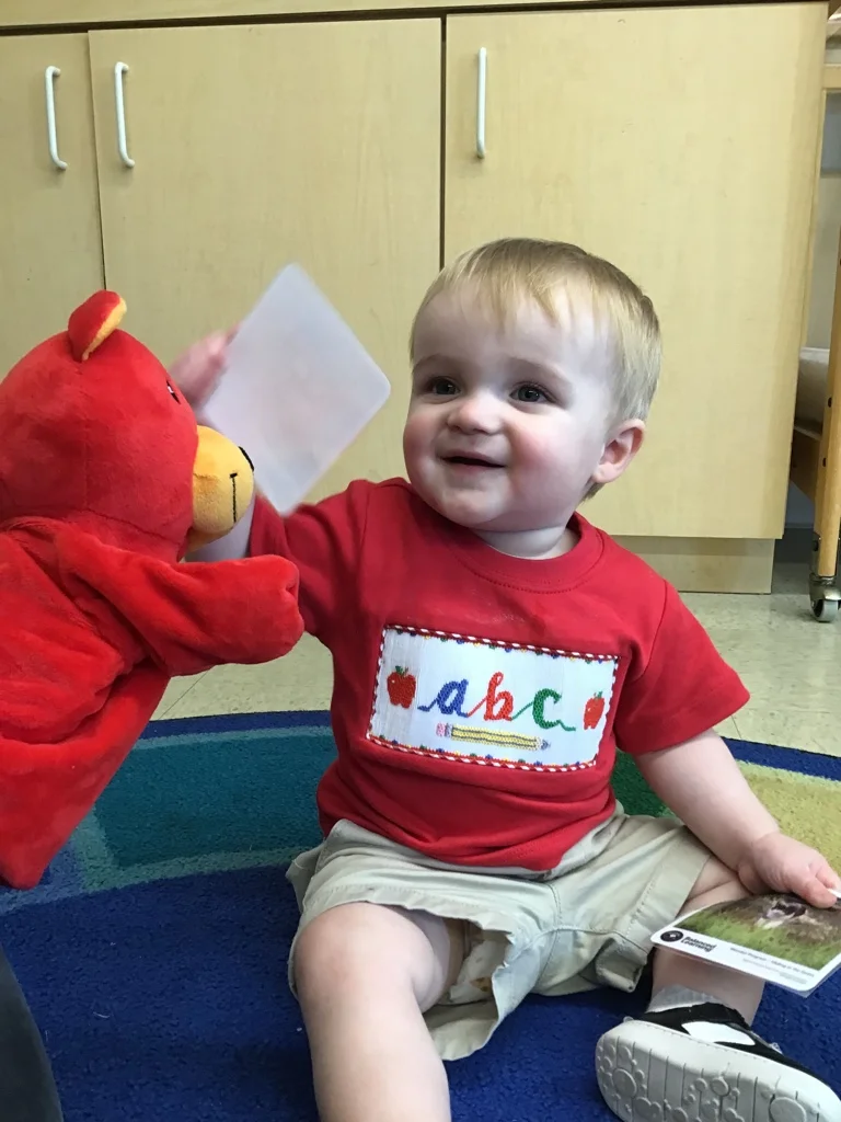 Toddler child wearing a red ABC shirt smiling holding a flash card with Benjamin the bear, a red stuffed bear puppet