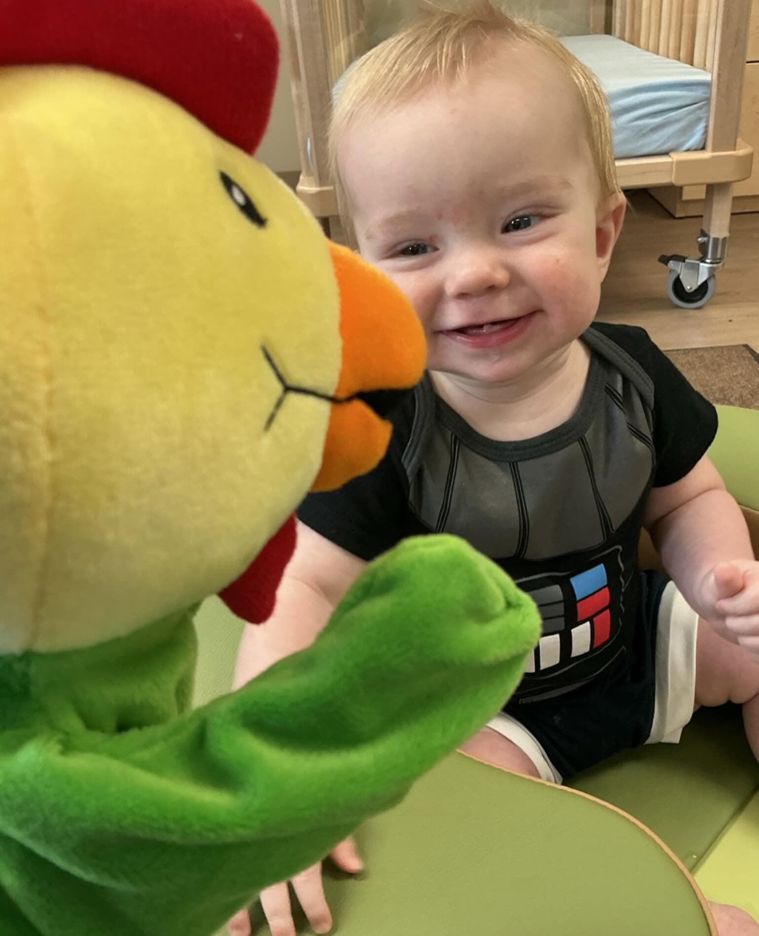 A smiling infant interacts with the Primrose Friend Percy the Rooster puppet during a music activity, building social, emotional, and early communication skills through songs and movement.