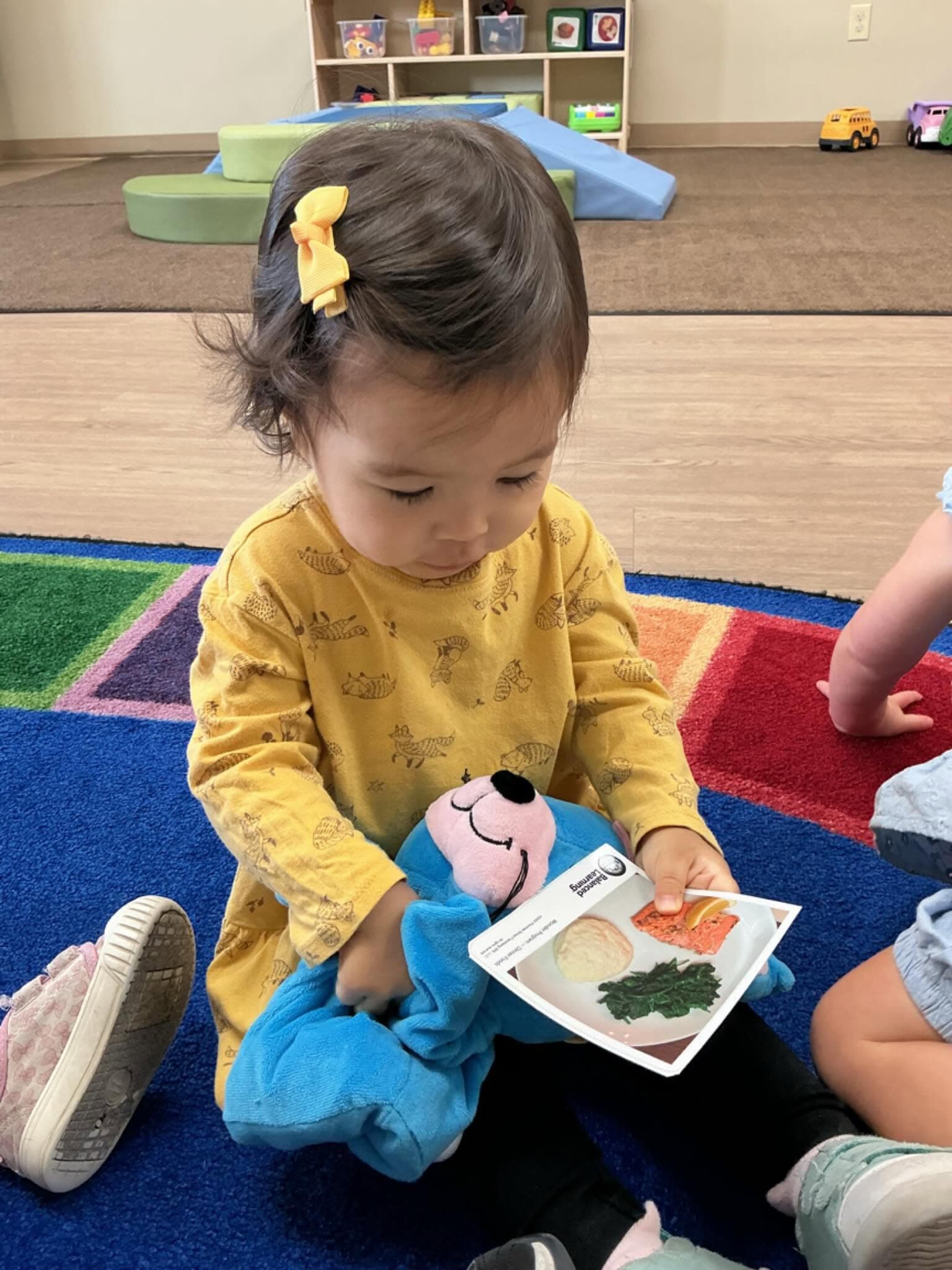 A toddler at Primrose School participates in a hands-on mealtime learning activity by holding a plush toy and a picture card of dinner foods while exploring pretend play, language development, and social skills with classmates.